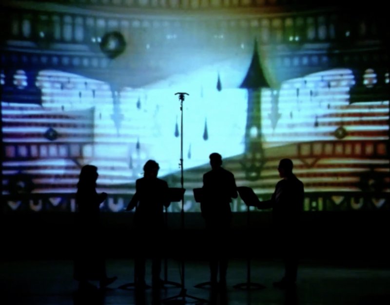 Silhouetted figures stand before a projected backdrop of blue and white architectural elements with dark accents. The backdrop features abstract shapes and geometric patterns.