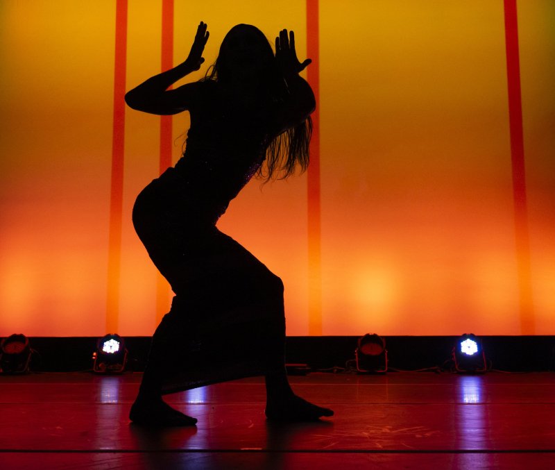 Silhouette of dancer with raised hands against orange backdrop with red stripes.