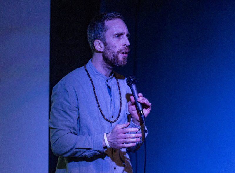 Man with beard in light blue shirt and beaded necklace holds a microphone, speaking against a dark blue background. Blue lighting illuminates the scene.