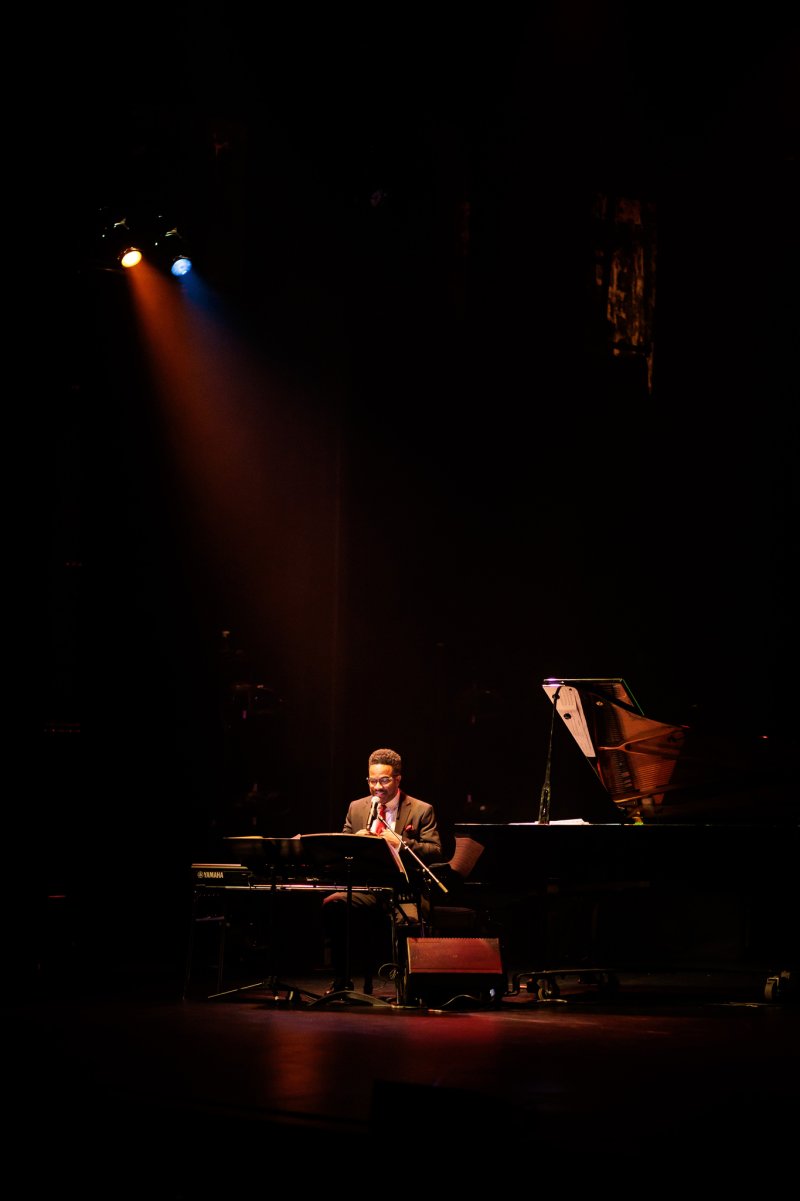Scott N Andrew's "Suite Life" (2022-2023) shows a musician in a dark suit playing an instrument on a stage, illuminated by orange and blue spotlights.