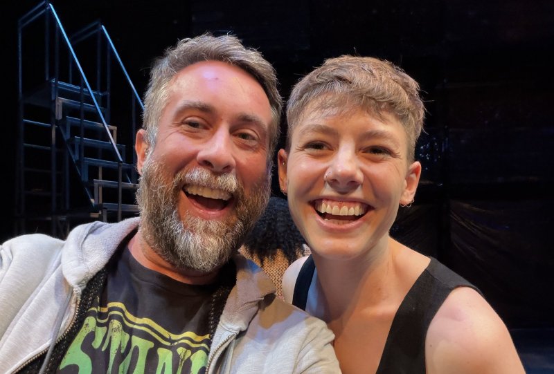 Close-up selfie of two smiling people, a bearded man and a short-haired woman, against a dark, blurred background of a stage set.