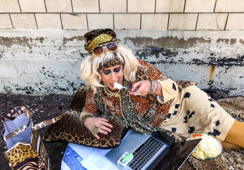 Drag queen in leopard print and blonde wig reclines outdoors, eating from a spoon while looking at a laptop.