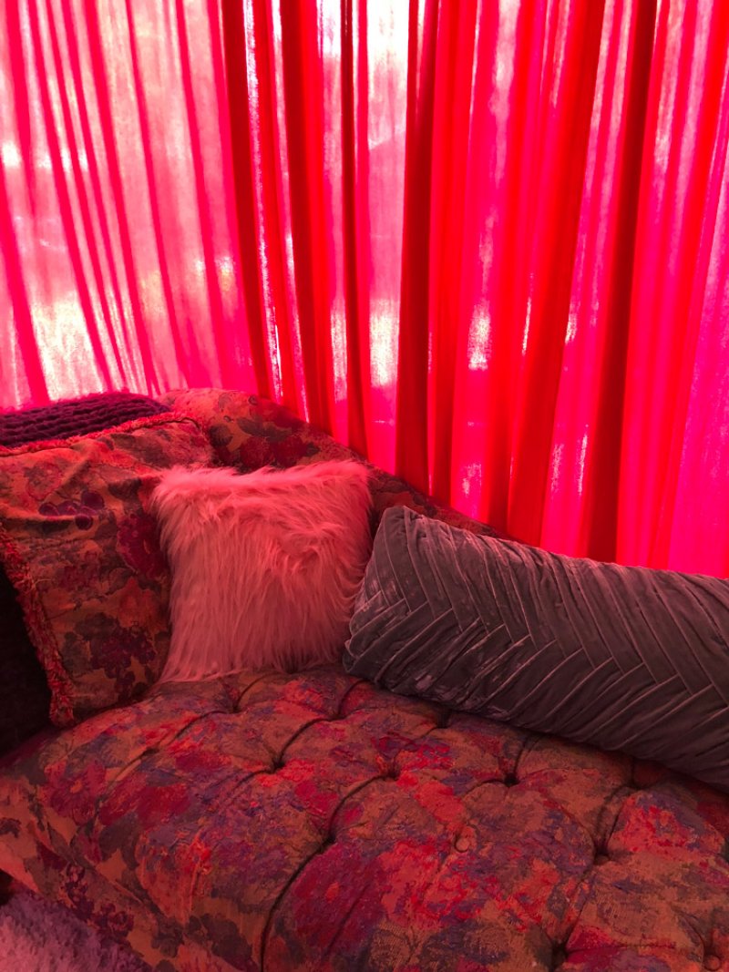 Vibrant pink curtains frame a chaise lounge with floral upholstery, a fluffy pink pillow, and a grey bolster pillow.