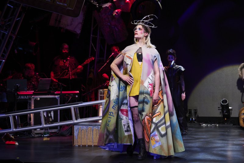 Blonde performer in a colorful, graffiti-covered cape and fishnets, standing on a dark stage with musicians and stage props in the background.
