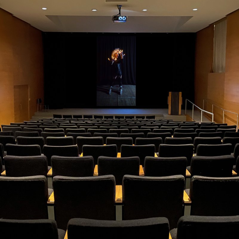 Auditorium with black seats facing a screen displaying a dark-haired figure in black, holding glowing orange flames.
