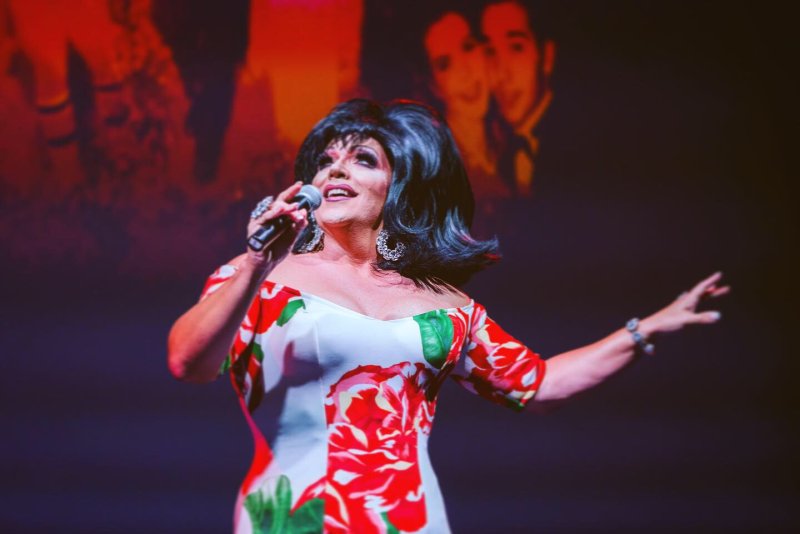 Drag queen in a white dress with red roses, black wig, and sparkling earrings sings into a microphone.