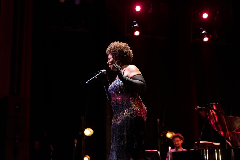 Singer with curly brown hair, in a shimmering dress and long gloves, holding a microphone on a dark stage with red lights.