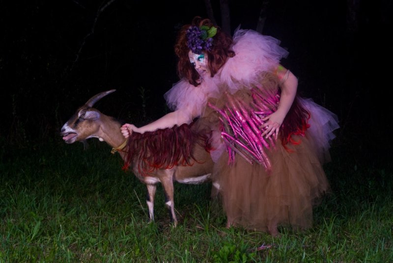 Drag performer in ruffled pink and brown dress with long pink claws, holding a goat with a red wig in a dark forest.