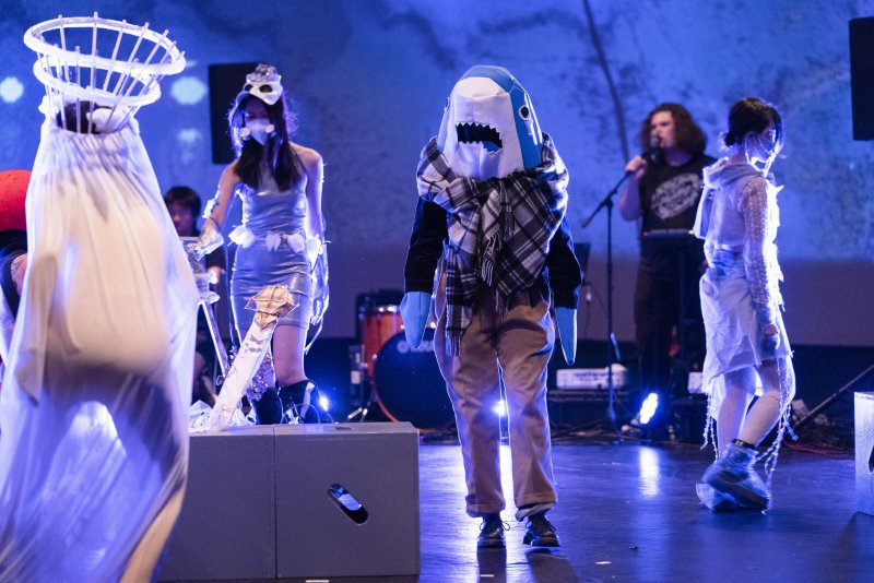 Performance art scene with figures in costumes under blue light. One wears a shark head and scarf, another a cage-like headpi