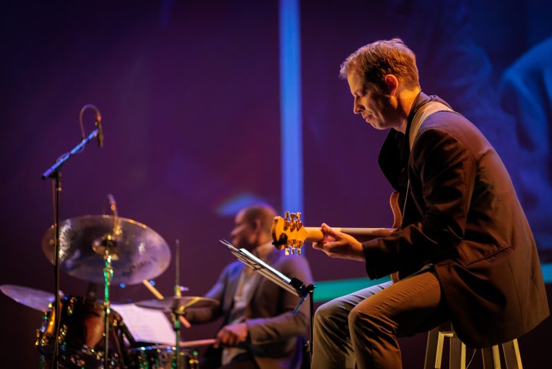 Man in dark suit playing guitar, side profile, with drummer blurred in background, stage lighting.
