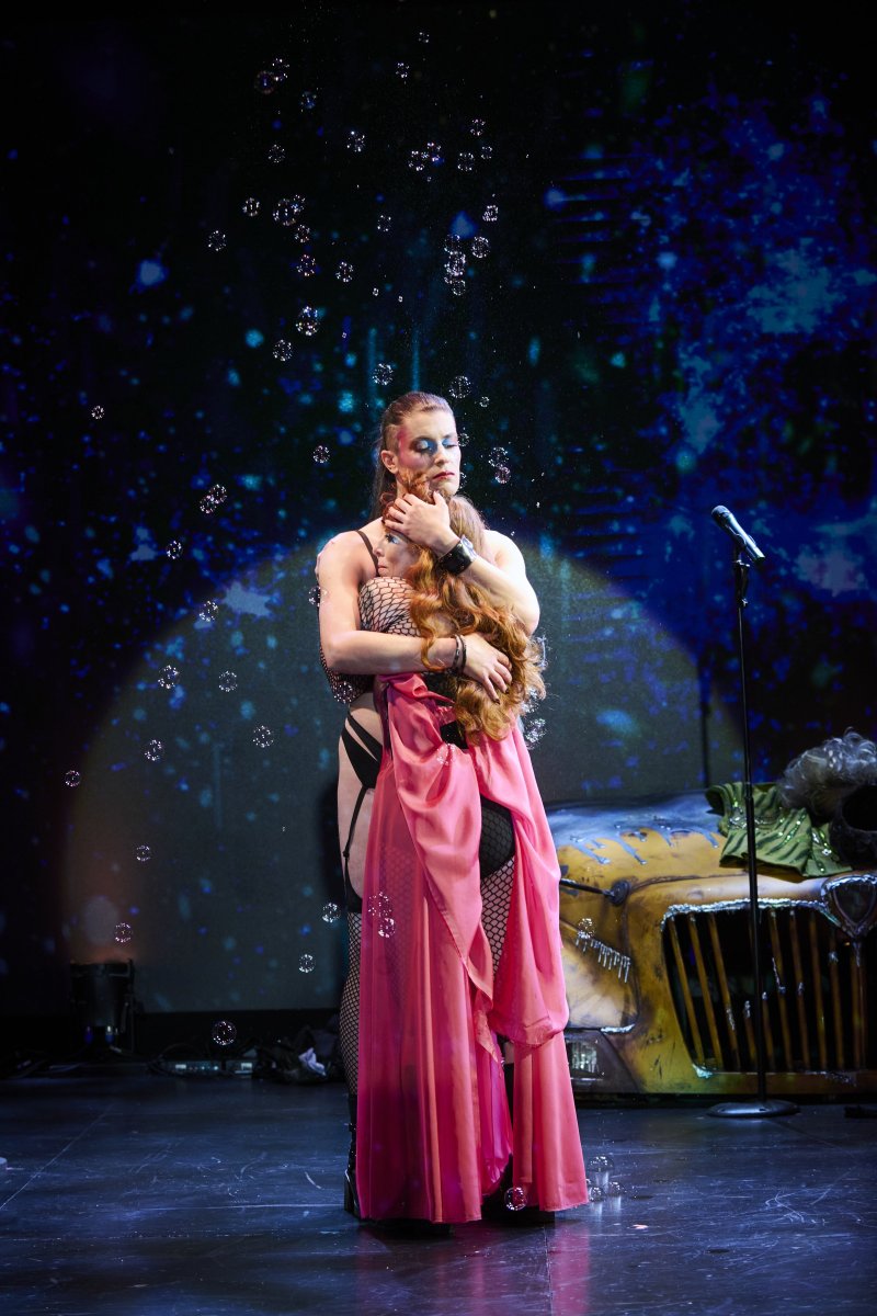 Two performers embrace on stage, one in a black bra and fishnets, the other in a flowing pink dress, with bubbles falling around them.