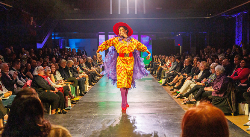 A model in a vibrant yellow and orange patterned dress, red hat, and purple boa poses on a runway before an audience.