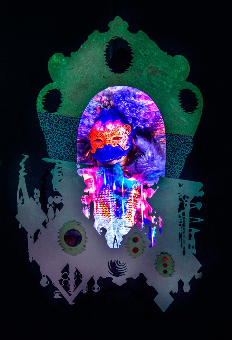 Mixed media artwork on black. Features a brightly lit, masked figure in an oval frame with purple and blue hues. Green and white abstract shapes frame the figure.