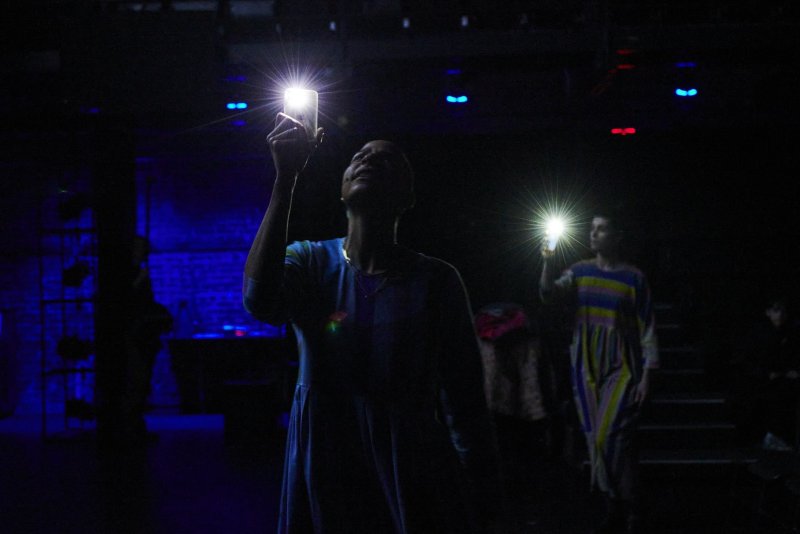 Two figures in a dark, theatrical space hold up bright cell phone lights. Blue and red stage lighting is visible in the background.