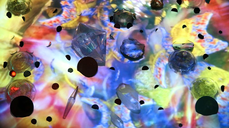 Abstract image of crystals, glass orbs, and dark circles on a vibrant, colorful, projected background.