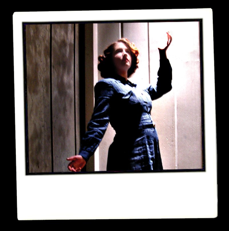 Woman in dark blue dress with red hair, looking up with arms outstretched, standing against a striped background.