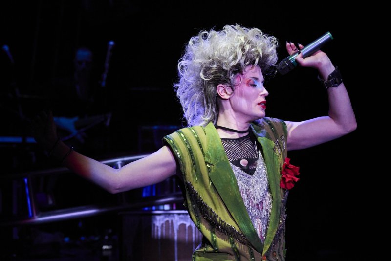 Performer with wild blonde hair, dramatic makeup, and a green vest sings into a microphone on a dark stage.