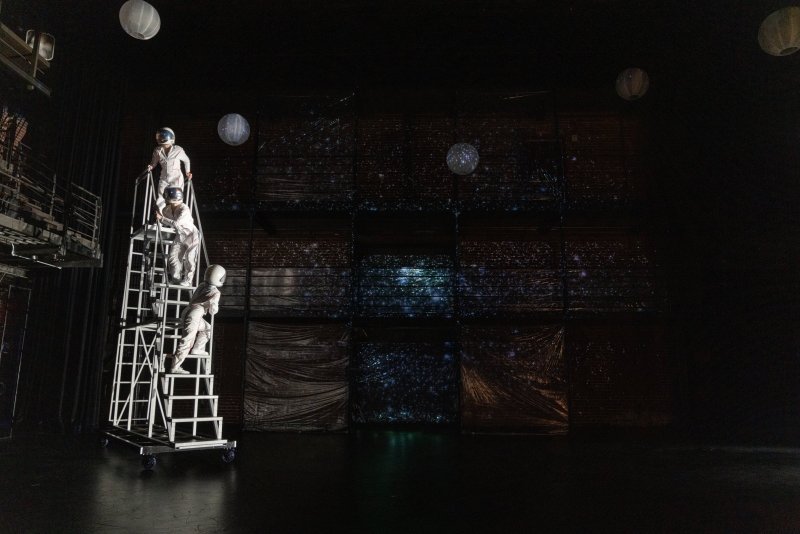 Three astronauts in white suits on a tall rolling ladder on a dark stage with glowing spherical lanterns and textured, starry backdrops.