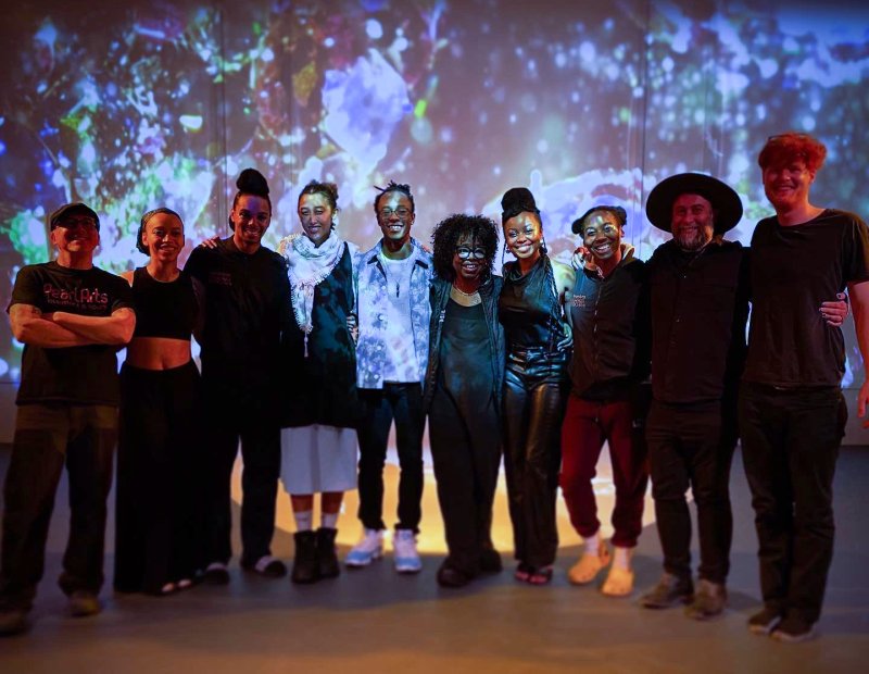 Group portrait of ten diverse individuals standing in a line against a backdrop of projected blue and white abstract shapes. The subjects are dressed in casual clothing, some with arms around each other.