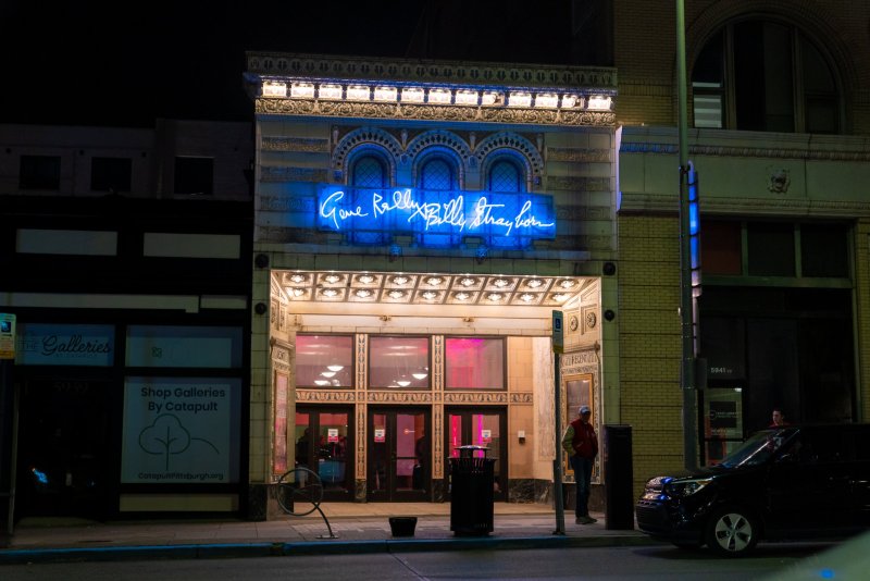 Night shot of an ornate building with "Gone Really Billy Strayhorn" in blue neon, pink light from inside, and a person on the sidewalk.