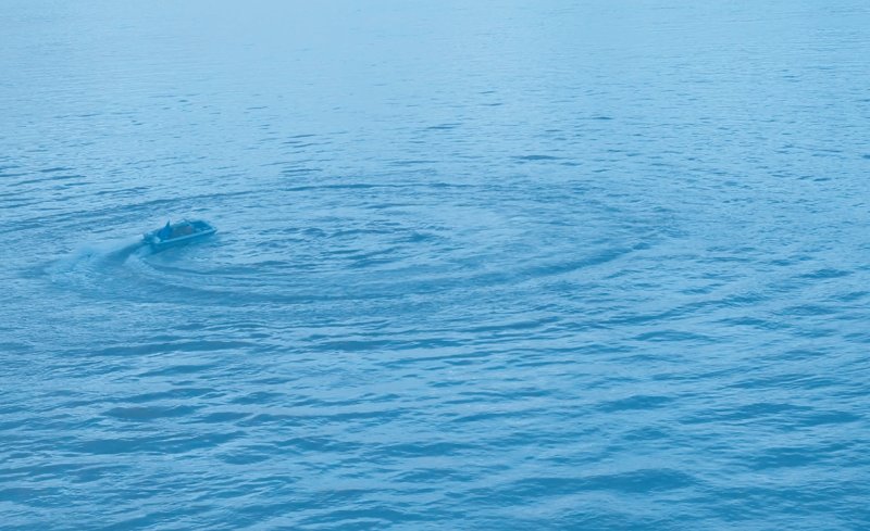 Small blue boat in the middle of a vast body of blue water, leaving circular ripples behind it.