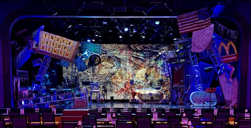 A theatrical stage set with a grungy, distressed backdrop, featuring a classic car, scattered props, and an American flag.