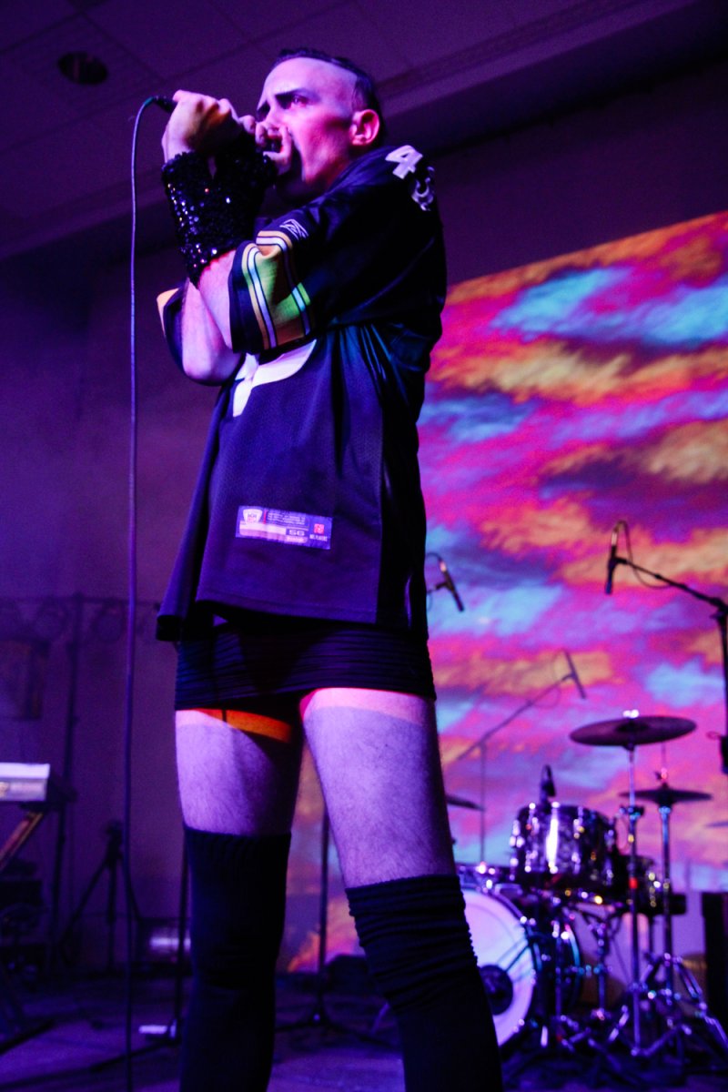 Performance shot in purple light: a singer in a black jersey and shorts, with black sequined wristbands, sings into a microphone. A drum kit and colorful light display are visible in the background.