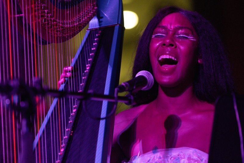 Performer with dark hair, glitter makeup, and open mouth stands next to a harp with red and purple strings, illuminated by pink and blue light. A microphone is in front of them.