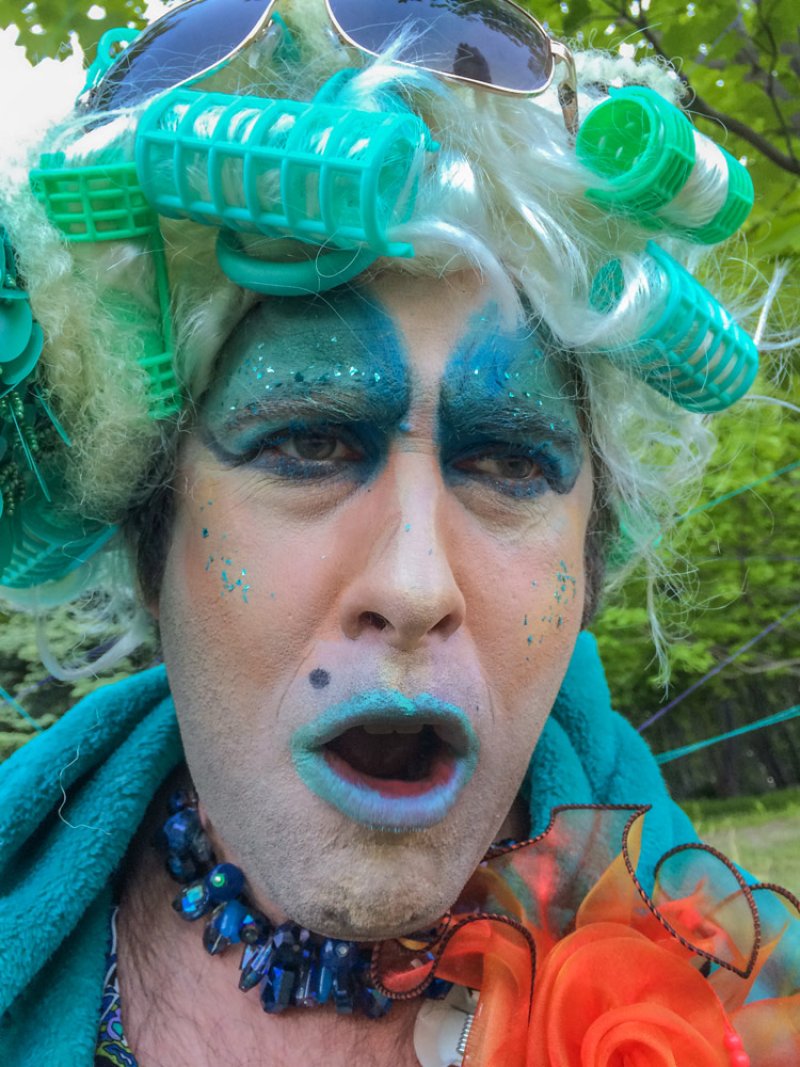 Close-up of a person in drag with blue eyeshadow, teal lipstick, and green hair rollers, looking at the viewer.