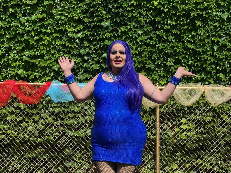 Drag queen with purple hair, blue dress, and fishnets, arms outstretched, in front of a chain-link fence and ivy-covered wall.