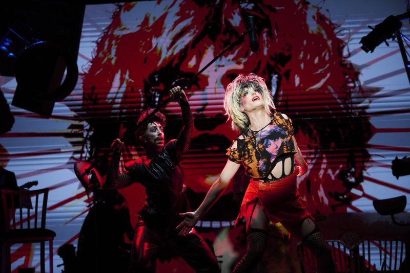 Two performers on stage, one in drag with a blonde wig and torn shirt, the other in black holding a mic stand, against a red graphic.