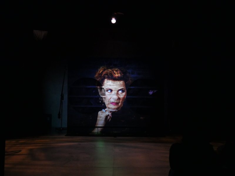 Projected image of an older woman with red hair, looking right, with a serious expression, on a dark stage.