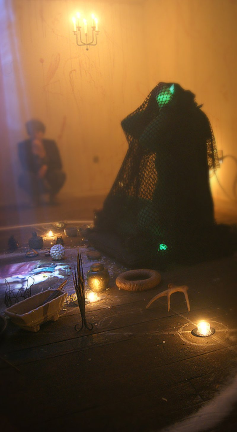 Warm-toned, hazy scene with a figure draped in dark netting, glowing green, surrounded by candles and ritual objects on a wooden floor. A blurred figure sits in the background, beneath a lit candelabra.