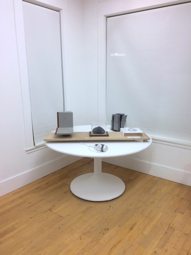 White room with a round white pedestal table displaying small, dark sculptural objects on a light wooden tray.