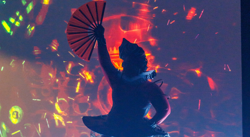 Dark silhouette of a costumed figure holding an open red fan aloft, against a vibrant red, orange, and yellow abstract light projection.