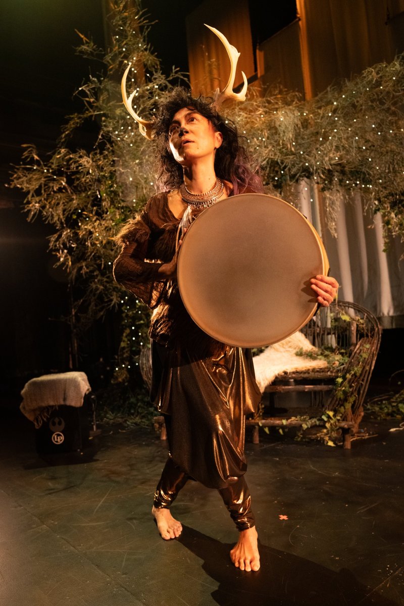 Barefoot person with antlers and dark, curly hair, wearing a gold outfit and holding a large drum, standing in a leafy, lit setting.