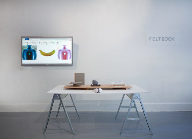 Gallery installation with a monitor displaying a blue vase, banana, and red vase, above a white table with objects.