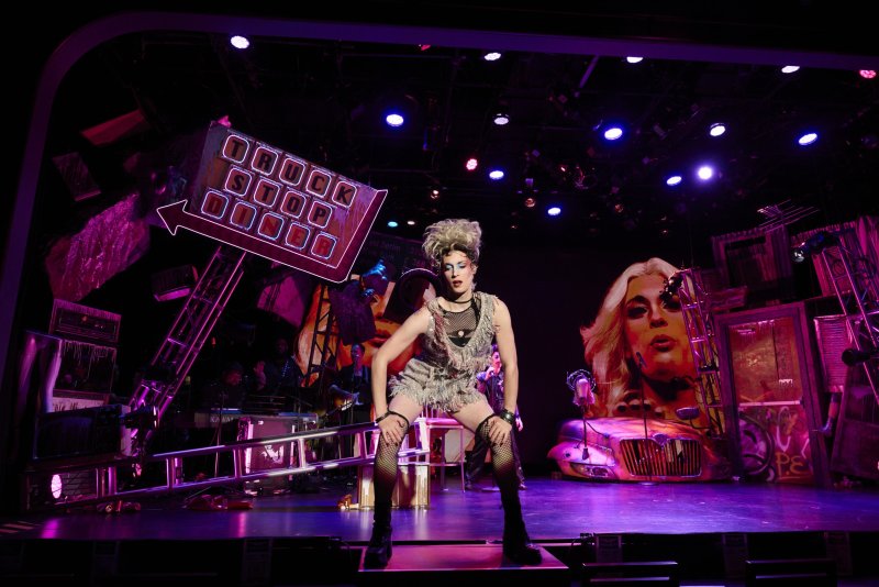 Drag queen in a blonde wig, fishnets, and a tattered dress on a stage with neon signs, a car, and a large face painting.