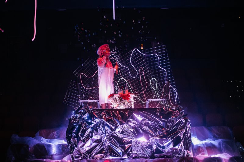 Performer in white, red hair, singing behind a shiny silver DJ booth with red flowers, neon grids, and bubbles in dark light.