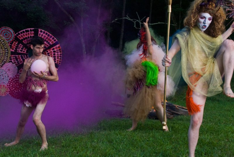 Three performers in elaborate costumes, one holding a pink orb, another a staff, amidst purple smoke outdoors.