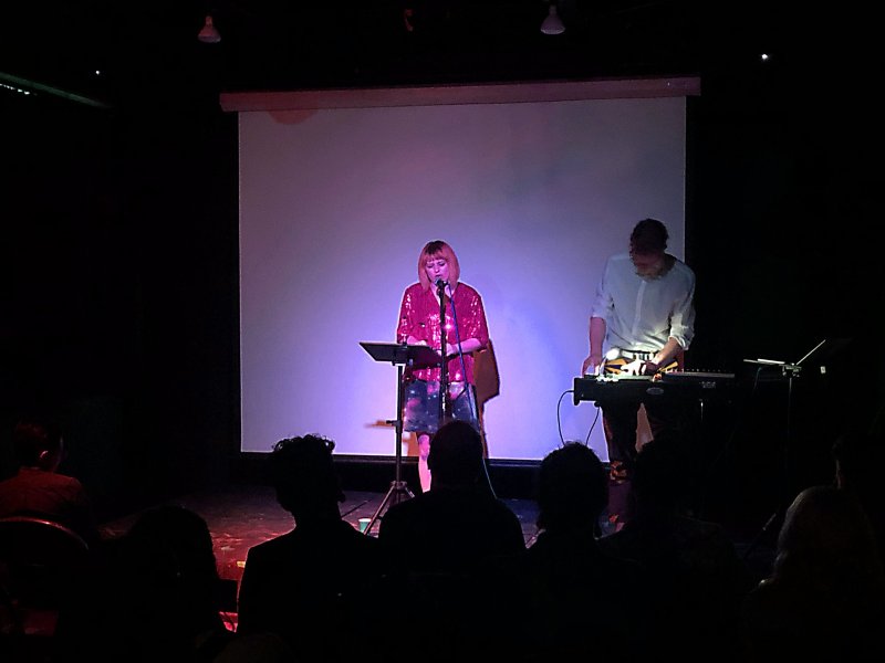 Dark stage view of two performers; one with pink hair at a mic stand, wearing a red sequin top and galaxy-print skirt, and one playing a keyboard. A white screen is behind them, with a silhouetted audience in the foreground.