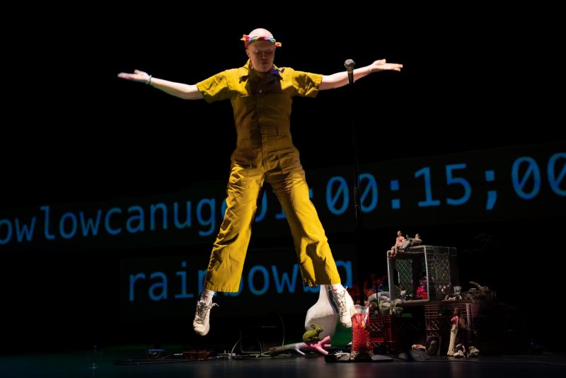 Performance art piece with a figure in a yellow jumpsuit mid-jump, arms outstretched, against a black backdrop. Blue text and a timer are projected behind them. Below, a collection of objects including crates, dolls, and plants are arranged.