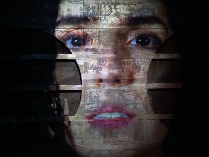 Close-up of a woman's face, distorted by horizontal black bars and a glitch effect, with wide eyes and parted lips.