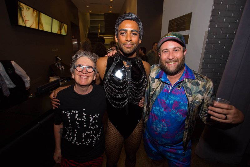 Three people pose indoors: one in a black bodysuit with chain detail, fishnet stockings, and silver glitter hair; one in a black t-shirt with white dots and red patterned shorts; and one in a blue floral shirt and jacket.