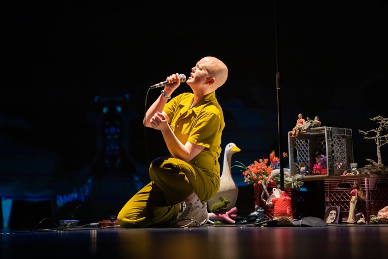 Performance shot: Bald person in yellow jumpsuit kneels, singing into a microphone. Stage cluttered with objects: goose, flowers, dollhouse, toys. Dark background with blue light.