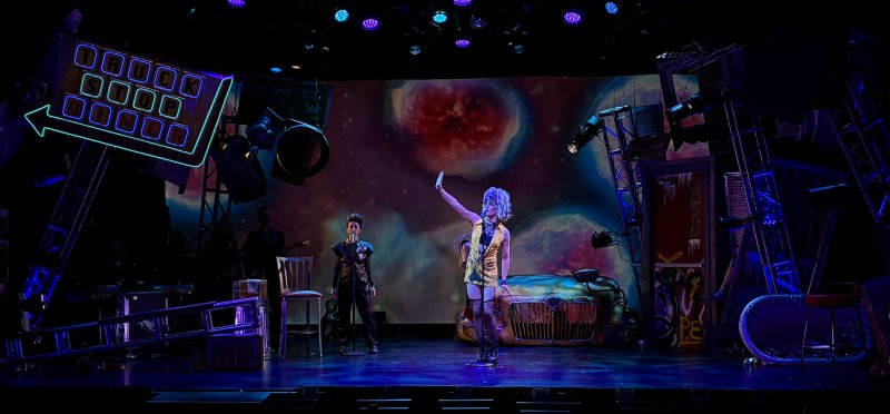 A theatrical stage with a performer in a yellow dress and wig, arm raised, beside another performer. Neon signs and a cosmic backdrop.