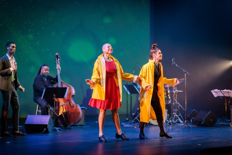 Two people in yellow raincoats dance on stage with a band, against a green and blue lit backdrop.