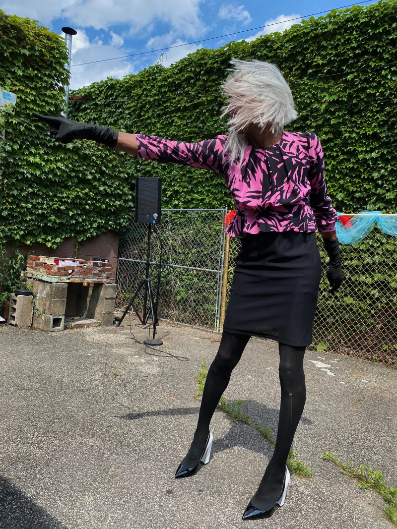 Person with white hair, pink and black patterned top, black skirt, and black gloves points right, standing on concrete.