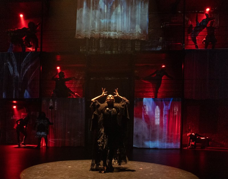Stage performance with a central figure in black raising hands, surrounded by silhouetted dancers on tiered platforms. Red lighting and projected gothic imagery create a dramatic, layered composition.