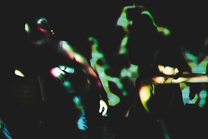 Two dancers in a dark room, illuminated by green and yellow projected light patterns on their bodies and the background wall.
